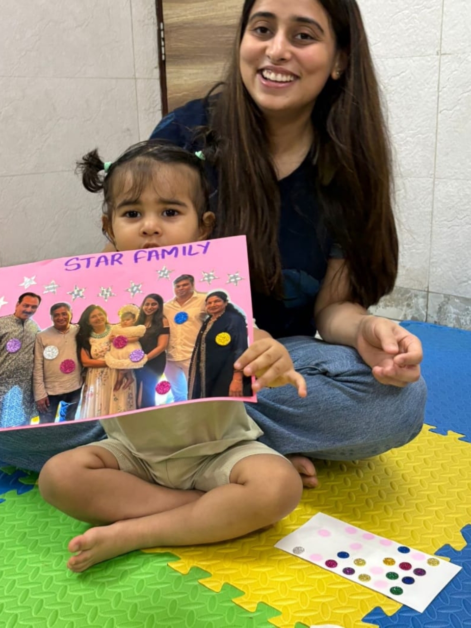 Child holding a colorful "Star Family" craft project while sitting with a smiling teacher.