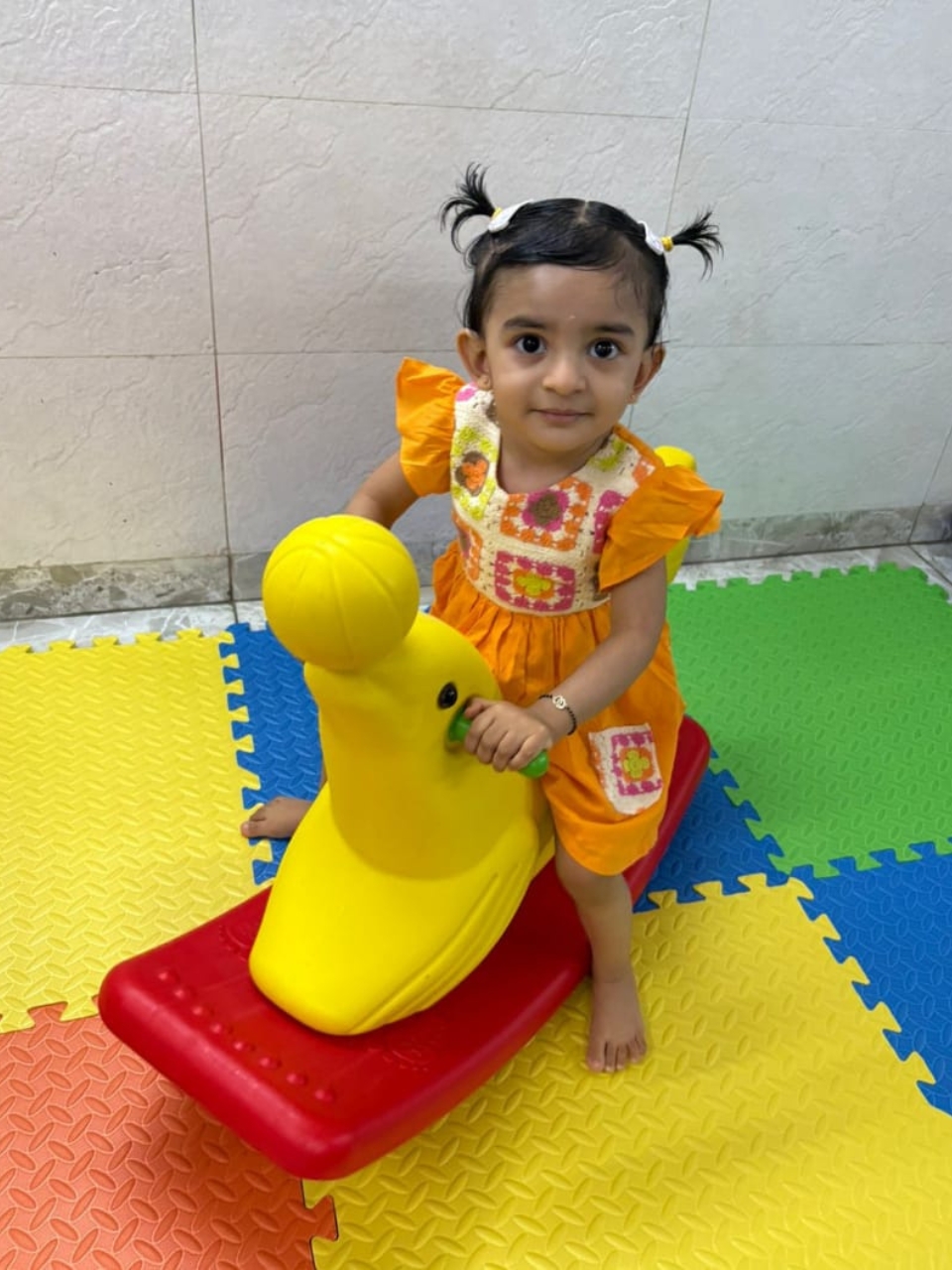 Smiling toddler sitting on a yellow rocking duck toy on colorful foam mats.