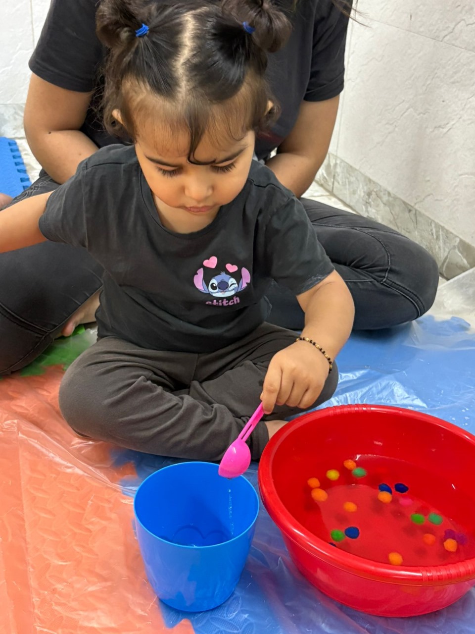 Little girl using a spoon to transfer colorful pom-poms from one bowl to another during a sensory activity.
