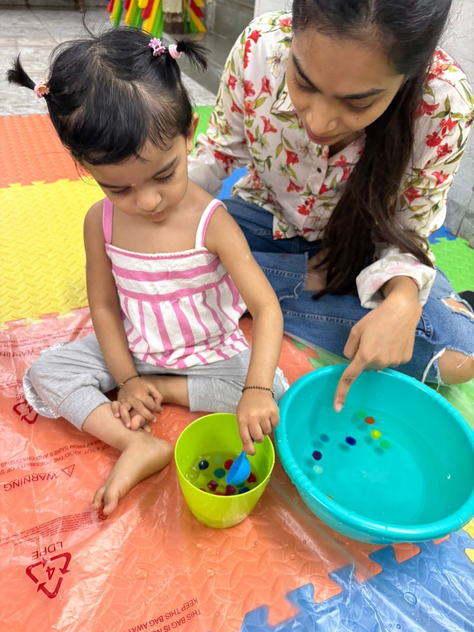 Toddler transferring pom-poms using a spoon during a water play activity with teacher assistance.