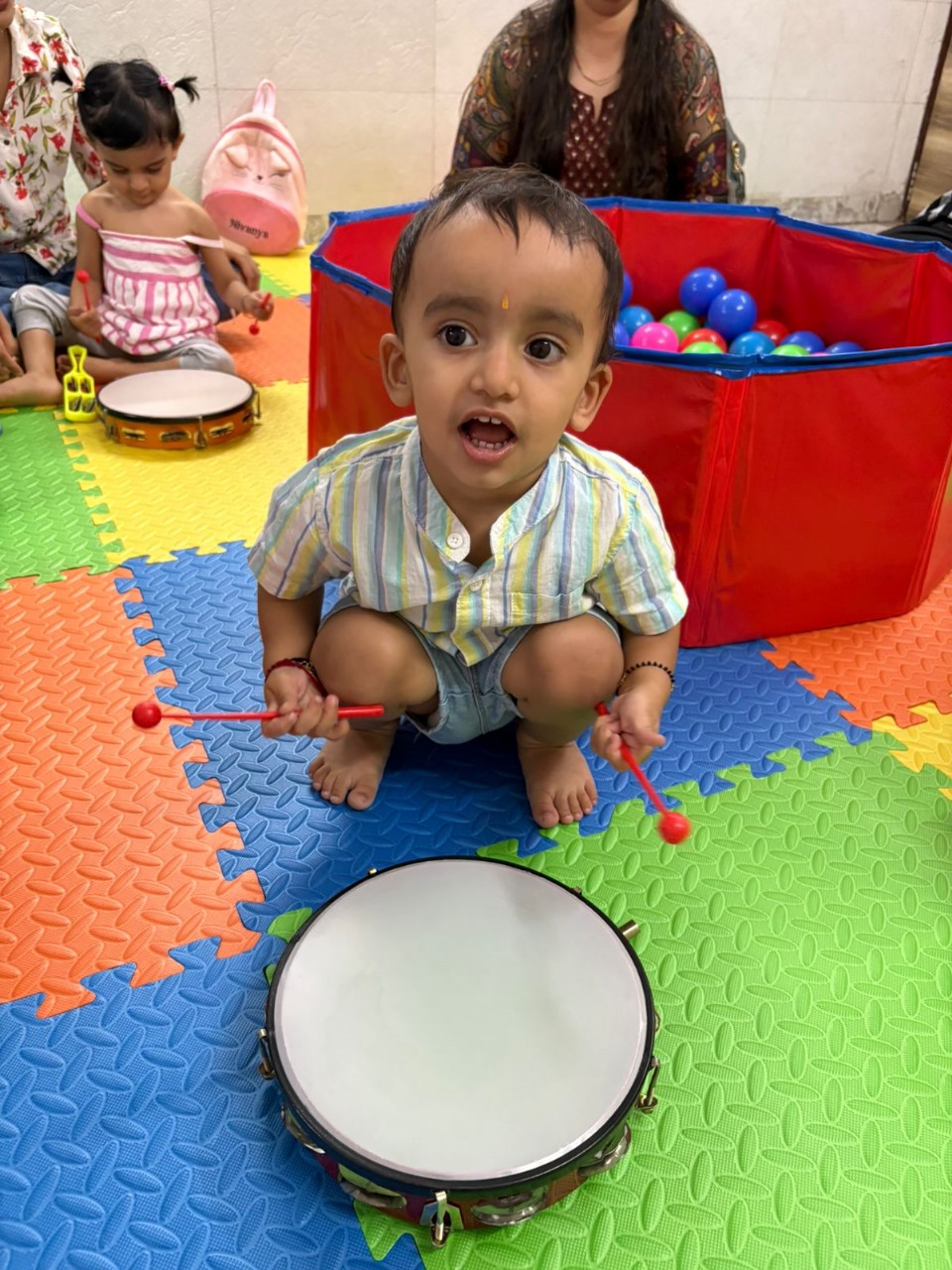 Toddler playing a toy drum with sticks during a fun music activity on a colorful play mat.