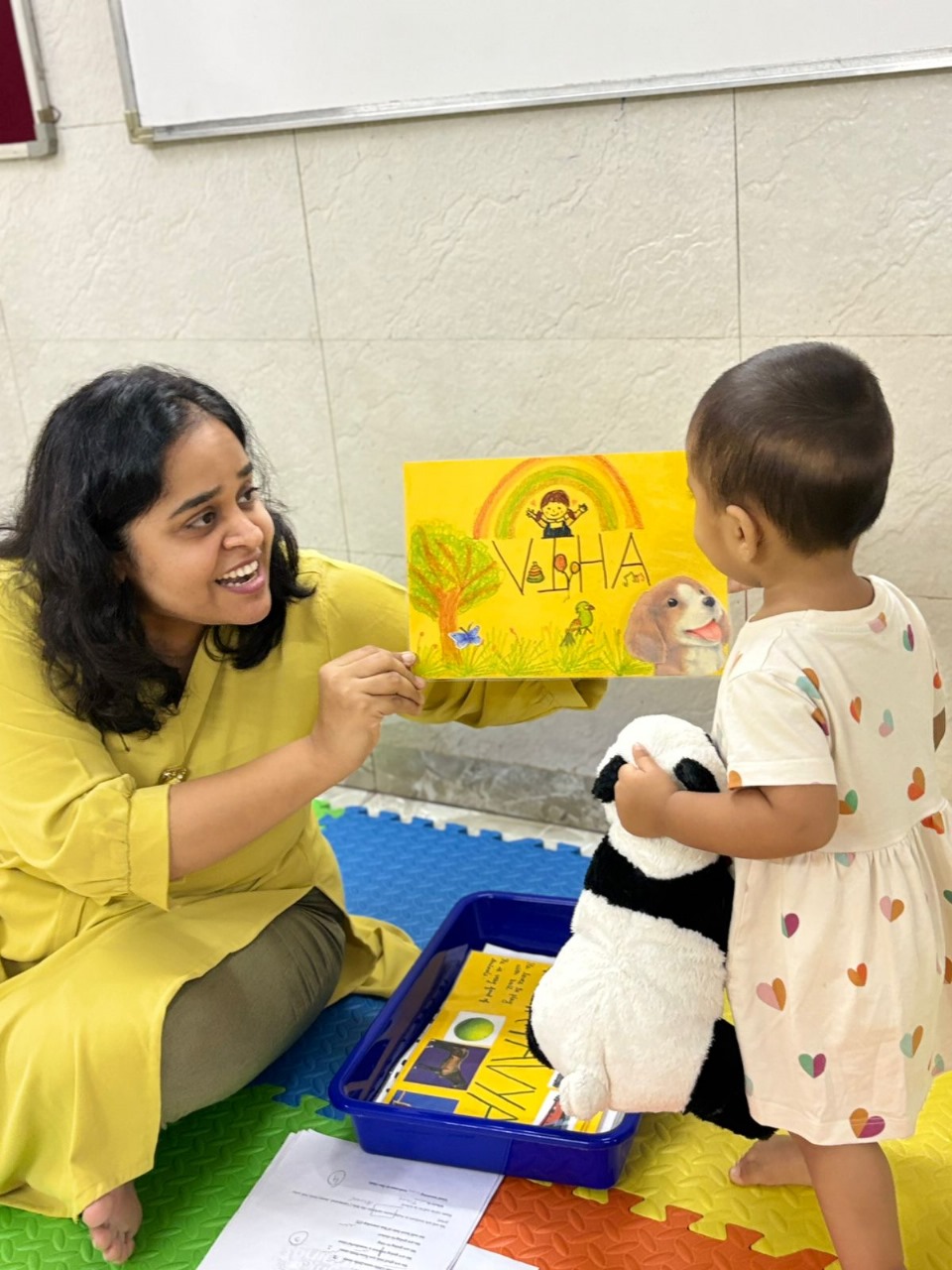 Teacher showing a colorful educational card to a toddler holding a stuffed toy.